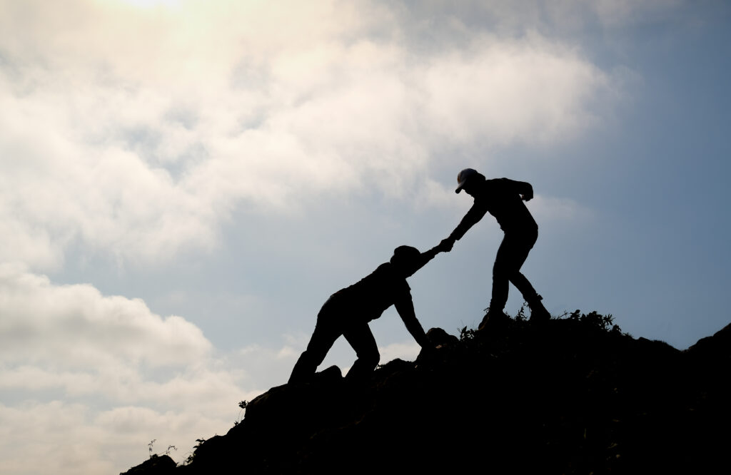 Silhouette image of one friend try to pull his other friend over the cliff of mountain and they climb and help each other during day time of travel.